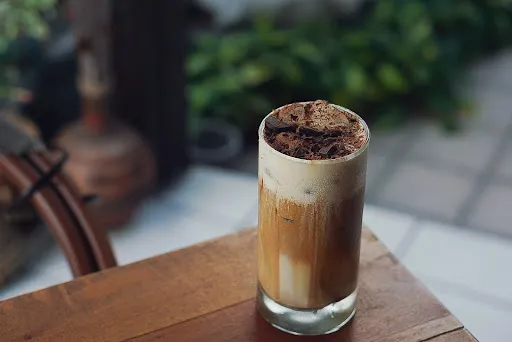 Layered iced coffee in a tall glass with cocoa dusting, blurred greenery in the background